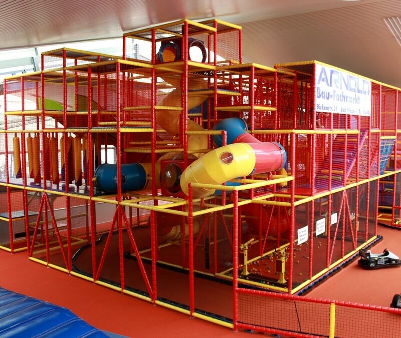 Stell dir ein großes Kletterlabyrinth vor, eine aufregende Spielarena voller Spaß und Abenteuer. Du siehst eine bunte Struktur aus Seilen, Netzen und Plattformen, die in verschiedenen Höhen und Richtungen angeordnet sind. Kinder klettern und balancieren geschickt durch das Labyrinth, während sie lachen und jubeln. Die Arena ist umgeben von grünen Bäumen und hat einen weichen, sicheren Boden. Eltern stehen am Rand, beobachten und ermutigen ihre Kinder. Die Atmosphäre ist voller Freude und Energie, perfekt für einen Tag voller Bewegung und Spiel.